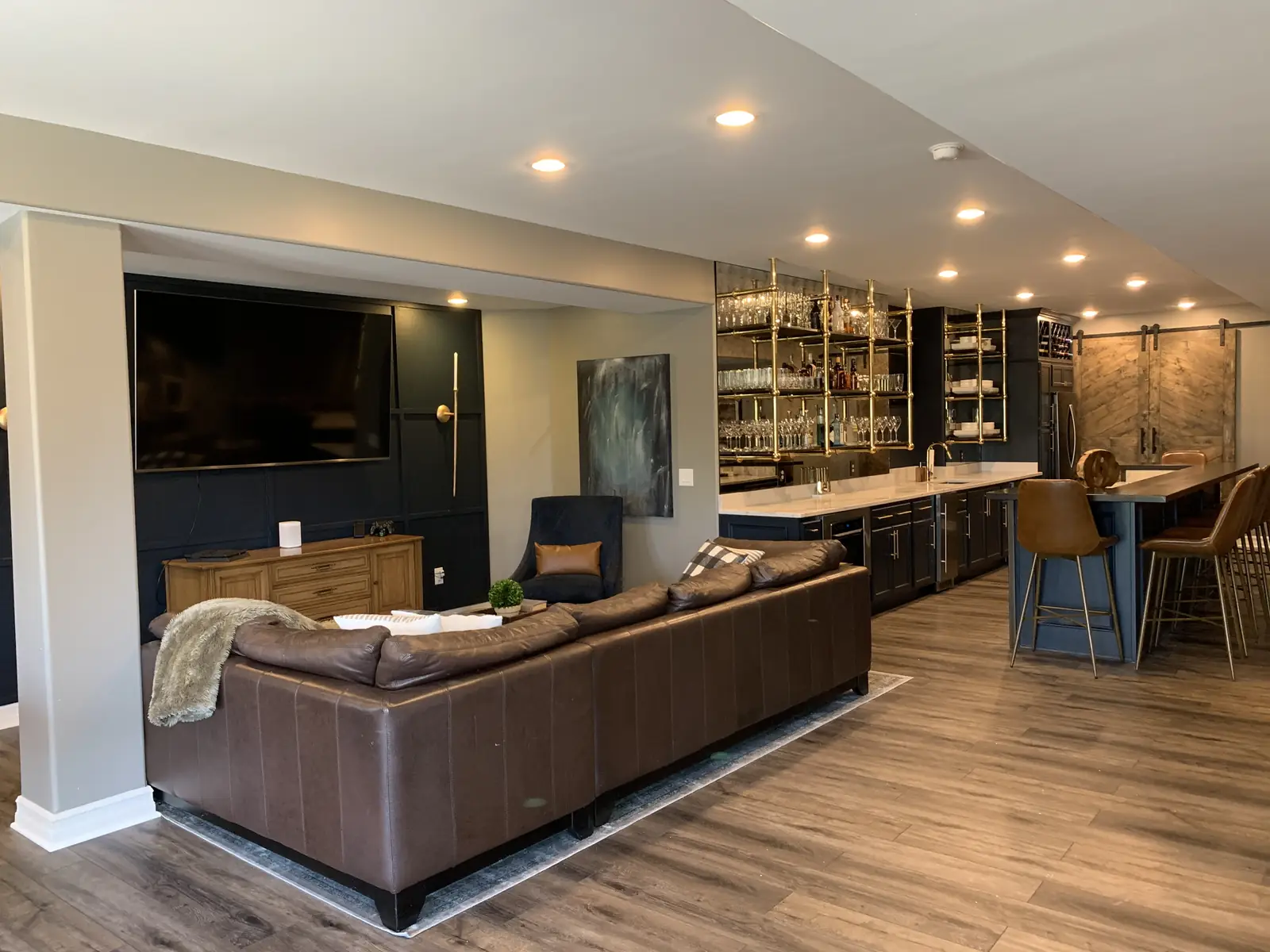 The Indian Spring lower level: a leather sectional facing a navy paneled TV wall, with the brass-shelved bar beyond