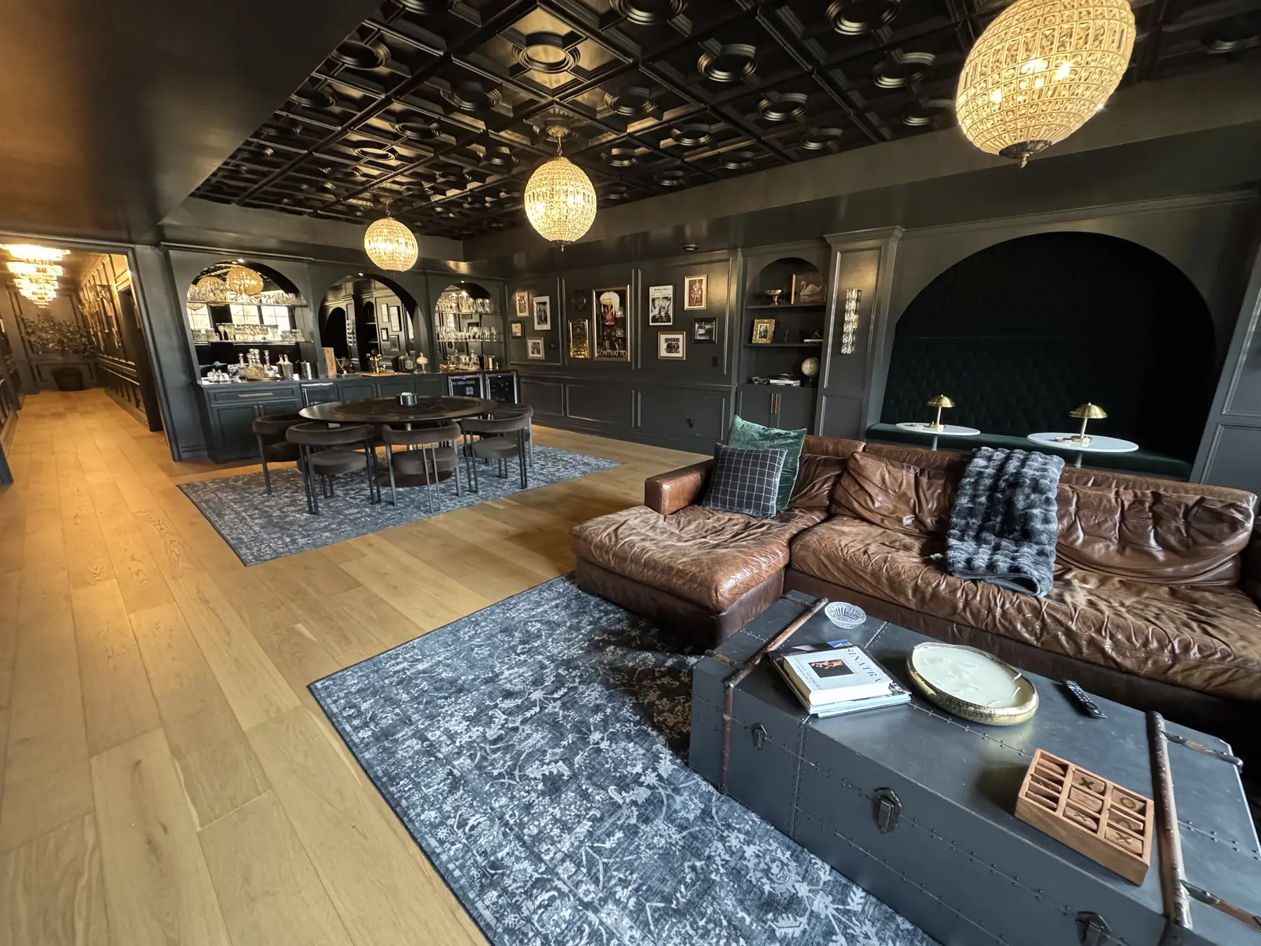 Dark green paneled speakeasy lounge with a coffered black ceiling, globe crystal chandeliers, a leather sectional and a bar